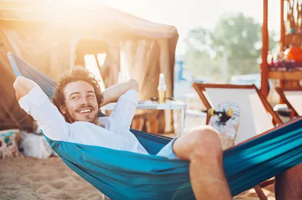 Ein lächelnder Mann entspannt in einer blauen Hängematte am Strand mit verschränkten Armen hinter dem Kopf, im Hintergrund ein Campingbus bei Sonnenschein.