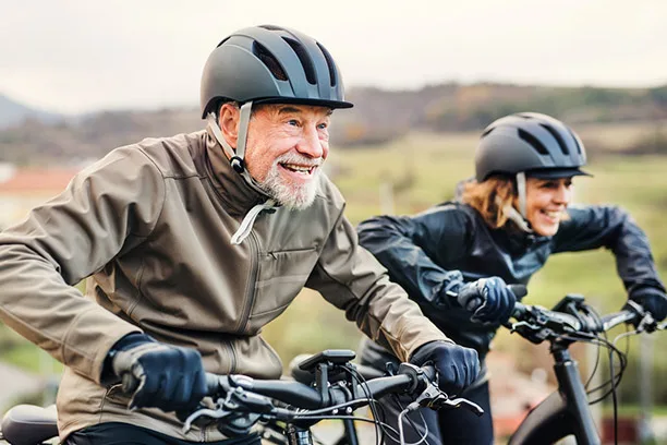 Ein älterer Mann und eine Frau fahren lächelnd mit Fahrrädern und Helmen durch eine ländliche Herbstlandschaft.