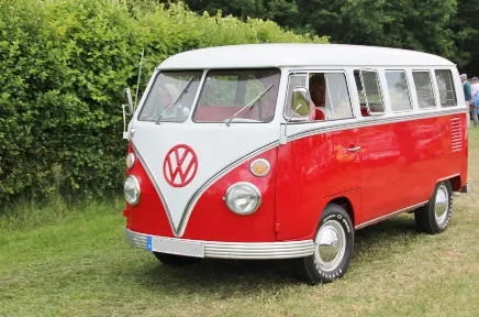 Ein rot-weißer VW Bulli T1 steht auf einer Wiese vor grünen Bäumen, das ikonische Volkswagen-Logo prangt auf der Front des klassischen Campingbusses.