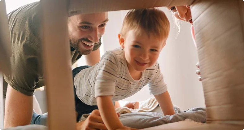 Ein lächelnder Vater und sein kleiner Sohn schauen gemeinsam fröhlich in einen Karton, umgeben von Umzugskartons in hellem Raum.