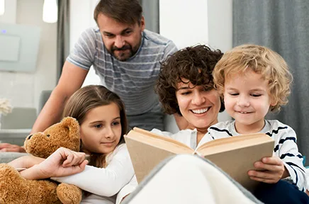 Familie liest gemeinsam ein Buch auf dem Sofa, während ein Mädchen mit Gipsarm und Teddybär zuschaut.