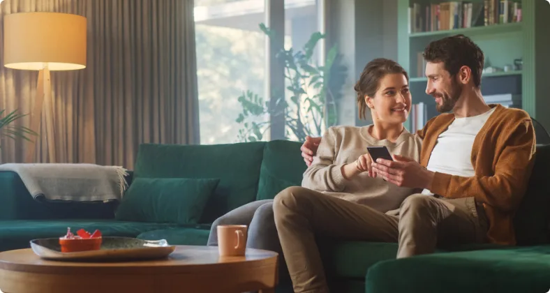 Ein Paar sitzt entspannt auf einem grünen Sofa in einem modern eingerichteten Wohnzimmer, schaut sich lächelnd an und hält ein Smartphone in der Hand.