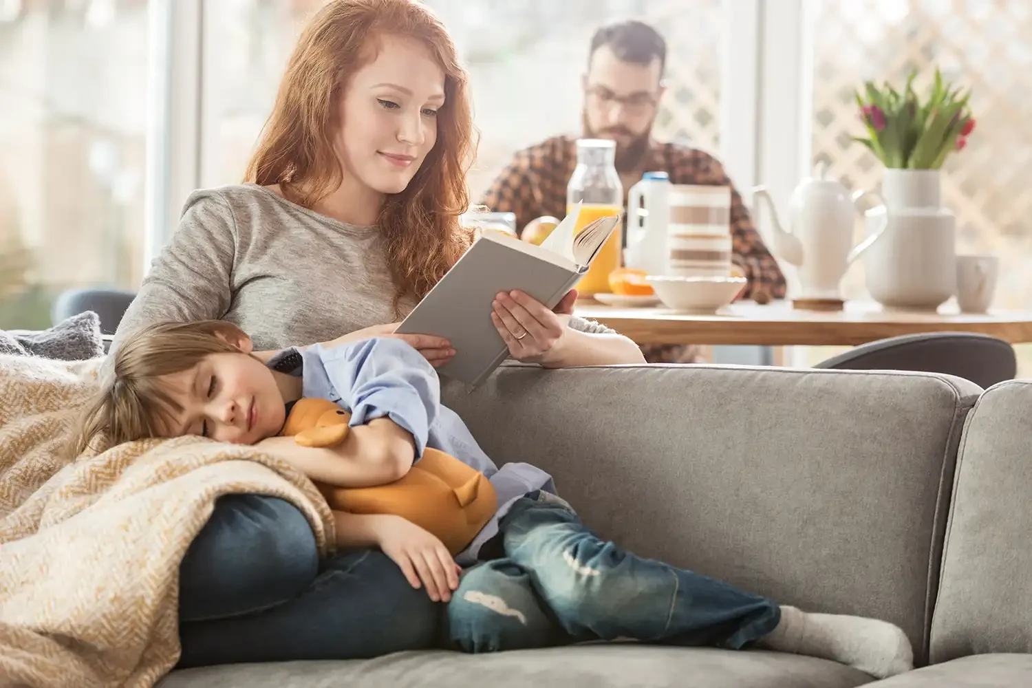 Eine Mutter liest ein Buch, während ihr Kind auf ihrem Schoß auf dem Sofa schläft, im Hintergrund sitzt der Vater am Esstisch.