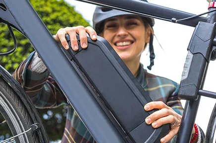 Lächelnde Frau mit Helm hält den Akku ihres E-Bikes in den Händen, im Hintergrund grüne Vegetation.
