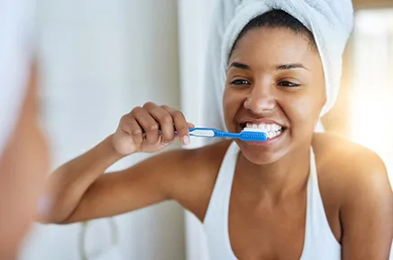 Junge Frau mit Handtuch auf dem Kopf putzt sich lächelnd die Zähne vor einem Spiegel im Badezimmer.
