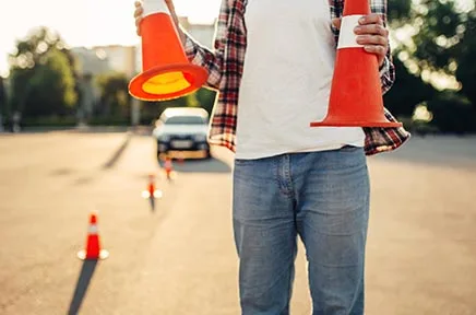 Person in Fahrschulausbildung hält Verkehrsleitkegel auf einer Straße mit Übungsparcours und Fahrschulauto im Hintergrund.