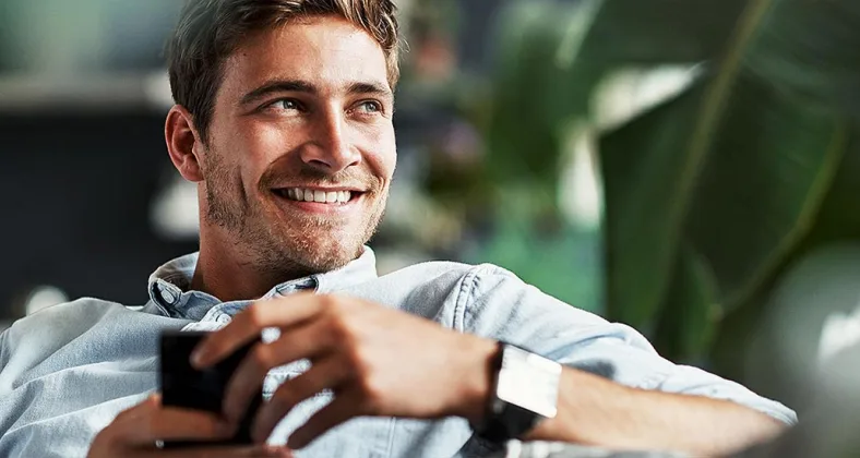 Ein lächelnder junger Mann mit braunem Haar sitzt entspannt auf einem Sofa, hält eine Tasse in den Händen und trägt ein graues Sweatshirt in einer hellen, grünen Umgebung.