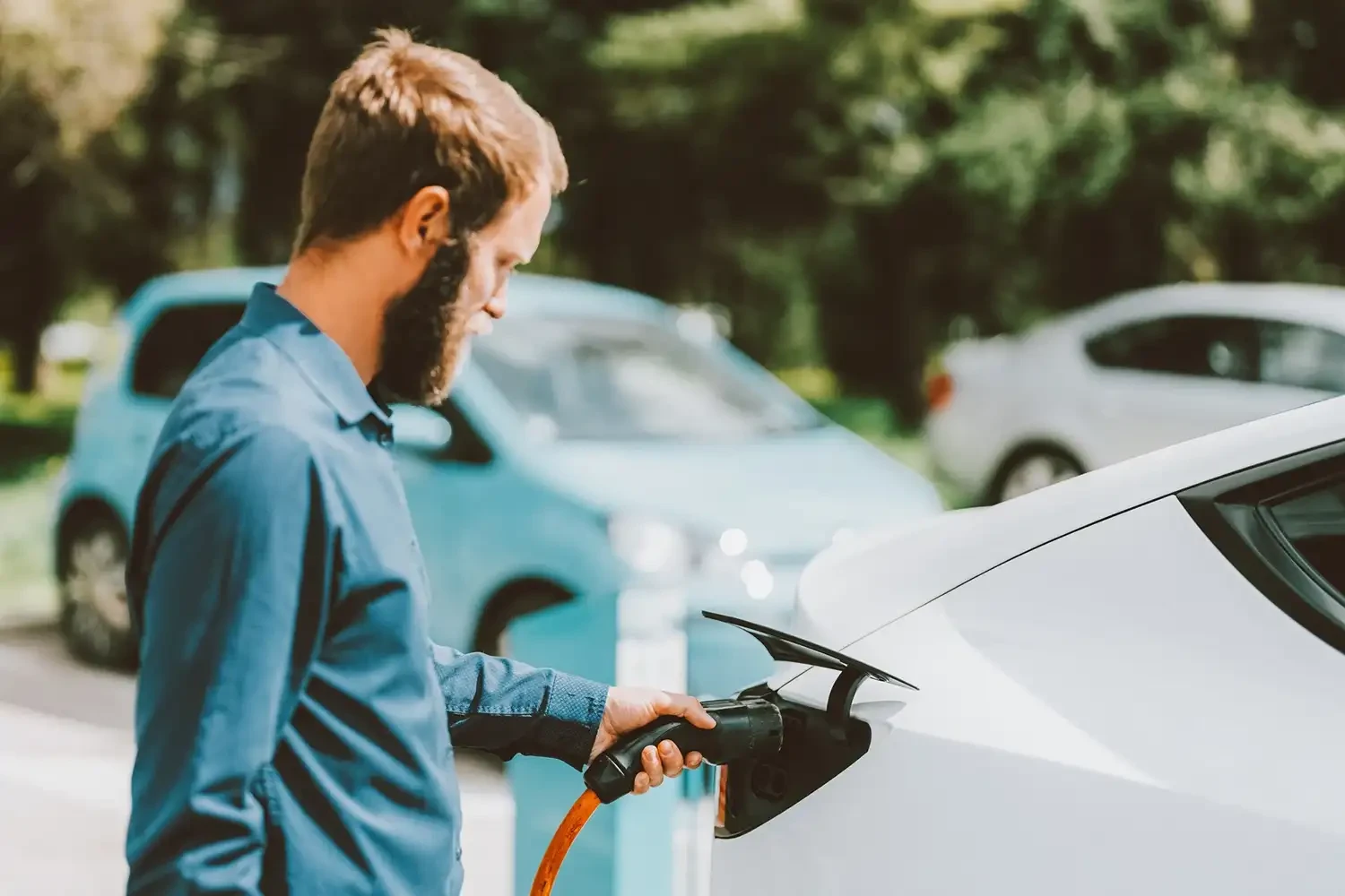 Ein Mann mit Bart lädt ein weißes Elektroauto an einer Ladestation auf, während im Hintergrund weitere Fahrzeuge auf einer grünen Straße zu sehen sind.