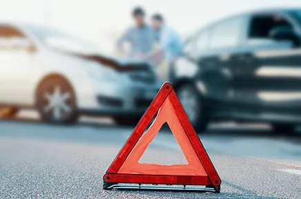 Warndreieck im Vordergrund nach einem Autounfall, im Hintergrund zwei beschädigte Fahrzeuge und zwei Personen im Gespräch auf der Straße.