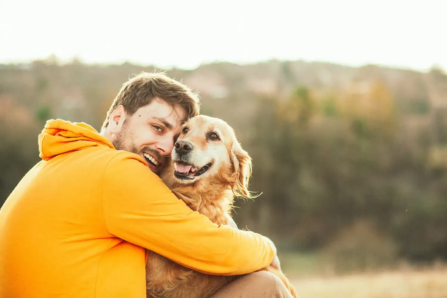 Person in orangefarbenem Hoodie umarmt lächelnden Golden Retriever vor unscharfem Naturhintergrund.
