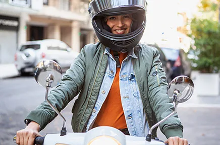 Lächelnde Person mit schwarzem Motorradhelm sitzt auf einem weißen Motorroller in einer Stadtstraße und trägt eine grüne Jacke über einer Jeansjacke.