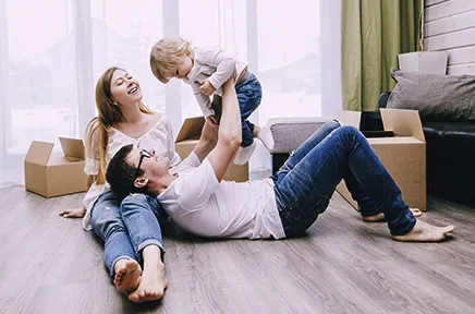 familie spielt gemeinsam auf dem boden: mutter liegt entspannt, vater hebt lachend ein kleinkind hoch, umgeben von umzugskartons in hellem wohnzimmer.