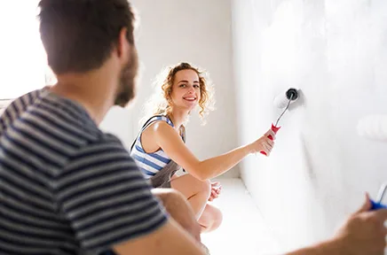 Junges Paar renoviert gemeinsam ihre Wohnung, die Frau streicht lächelnd die weiße Wand mit einer Farbrolle, während der Mann im gestreiften Shirt zuschaut.